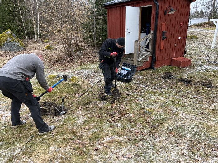 Två personer arbetar utomhus med att installera el till en vattenpump vid ett rött pumphus. De använder verktyg för att slå ner ett jordspett.