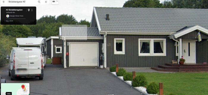 Grått hus med garage och vita fönster, vy från uppfart med parkerad vit skåpbil. Grön häck och trädgård, svensk flagga vid entrén. Google Maps etikett synlig.