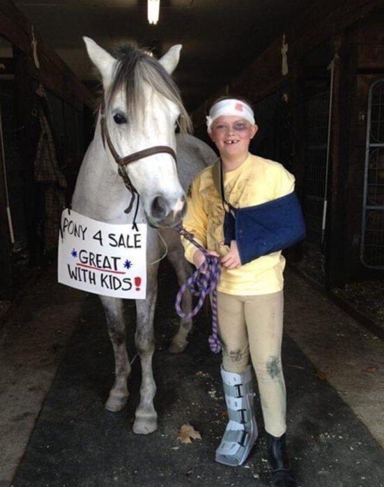 En pojke i gult med gipsad arm och ben står bredvid en häst som har en skylt "Pony 4 Sale, Great with Kids".