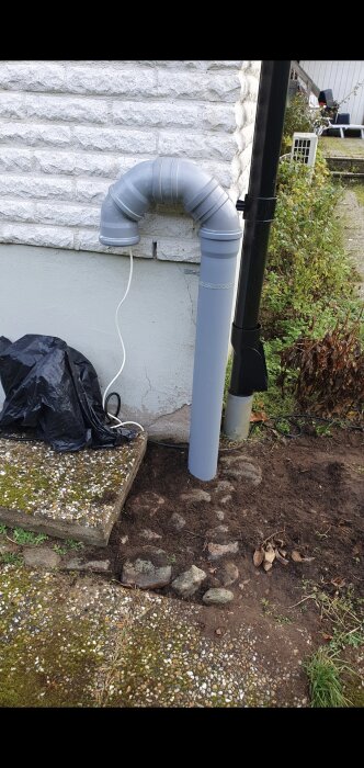 Grå avluftningsrör och fläkt installerade vid husvägg för att reducera radon, med svart kabel och plastpåse synliga bredvid.