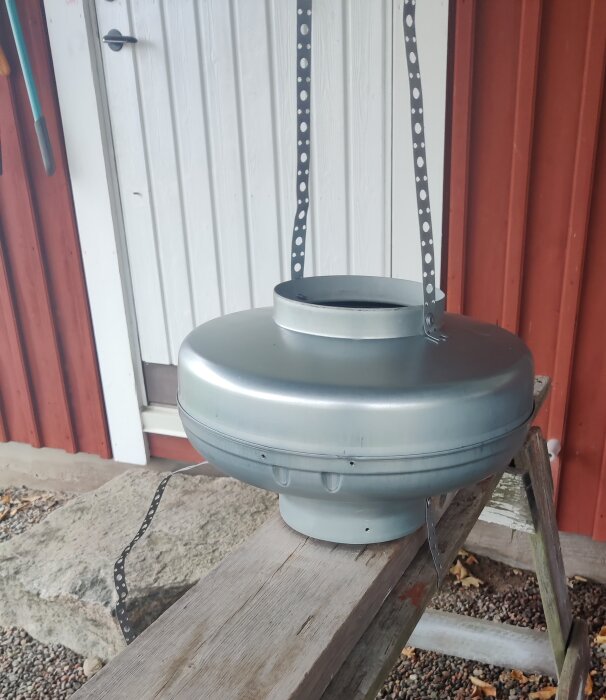 Metallfläkt fastsatt på en träbock utomhus, med hålband fästa i fläktens övre del, mot en röd och vit byggnadsvägg i bakgrunden.