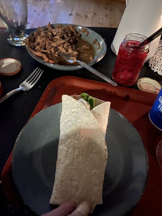 Tortilla fylld med pulled pork på tallrik, omgiven av skål med kött och glasburk med inlagd rödlök på bord.