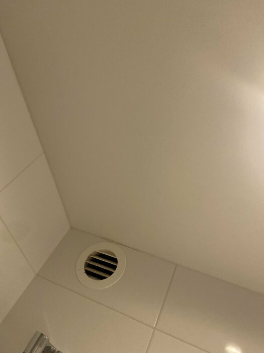 Vit ventilationsöppning i hörnet av ett badrum med vita kakelväggar.