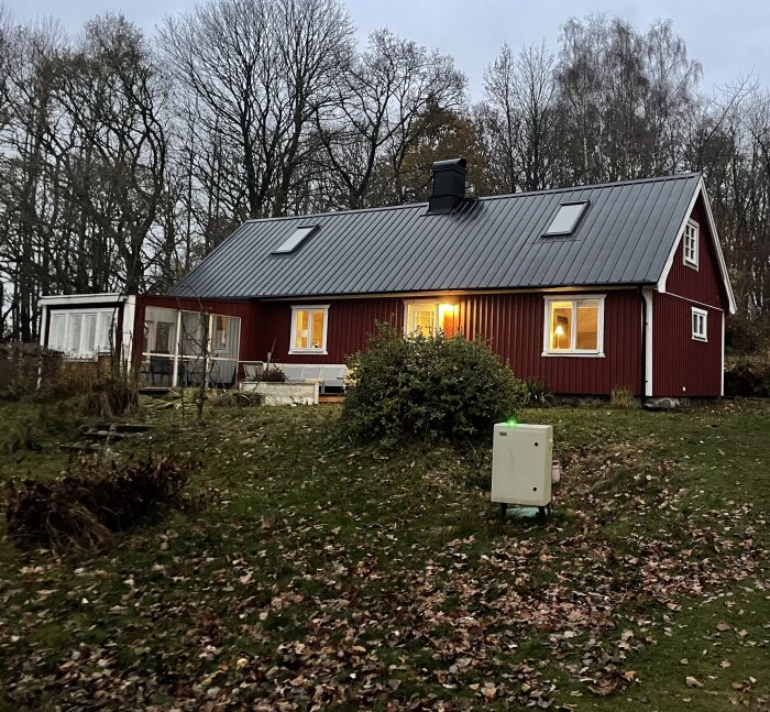 Elektronikskåp framför ett rött hus med grått tak omgiven av träd och gräsmatta, med slamavskiljarens brunnslock bakom.