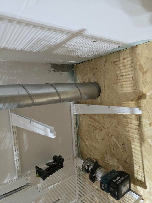 Ventilationsrör genom OSB-vägg med fräsverktyg och hyllplan, förberett för att installera duschens ventilation i förrådet.