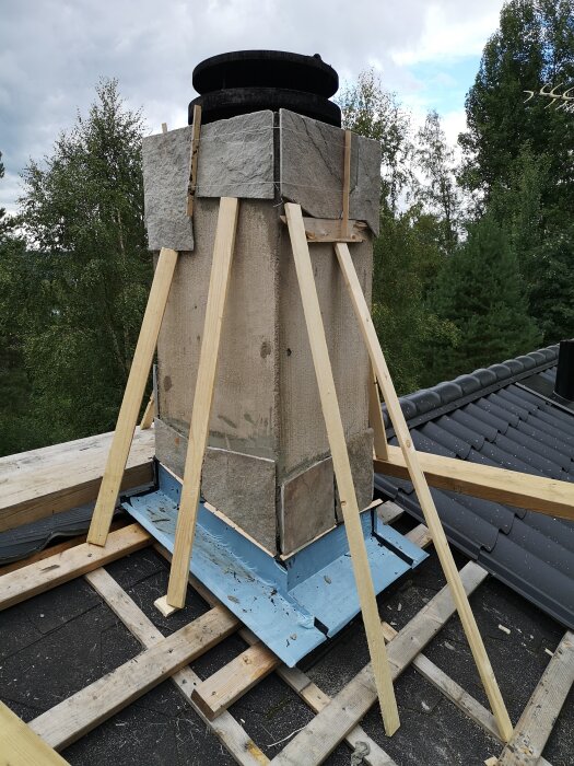 Chimney under renovation with wooden supports on a roof, visible chimney cap and surrounding trees in the background.