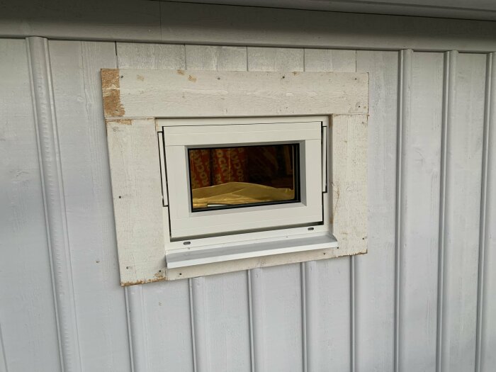 Nyinstallerat fönster med vit karm på garagets övervåning, omgärdat av olackerade träplattor mot en ljusgrå träpanel.