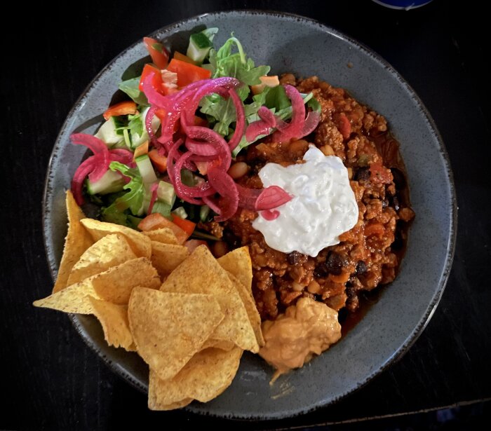 Tallrik med chili con carne toppad med gräddfil, sallad med picklad rödlök och nachochips.