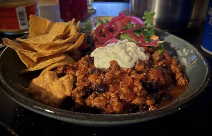 Tallrik med chili con carne med svarta bönor, gräddfil på toppen, picklad rödlök och sallad vid sidan, samt nachochips och ostsås.