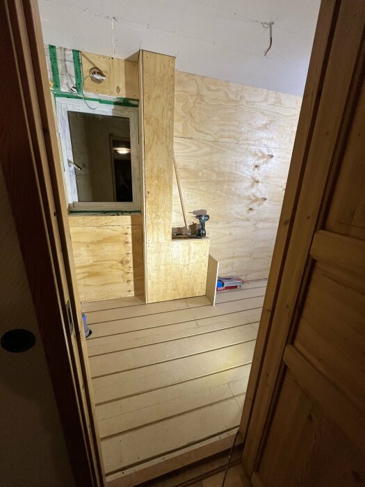 Rum under renovering med golvplattor och plywood på väggar, gipstak; syns tvättnedkastets ytterhölje i plywood, verktyg på golvet.