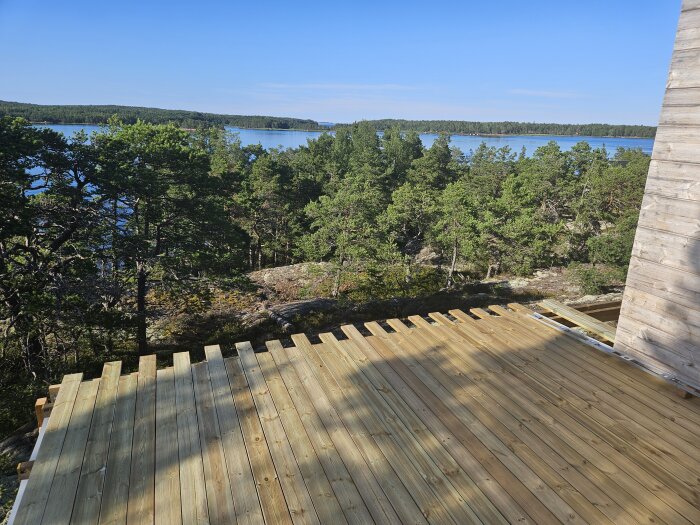 Träterrass med utsikt över en sjö och skog, del av ett byggprojekt. En trävägg syns till höger i bilden.