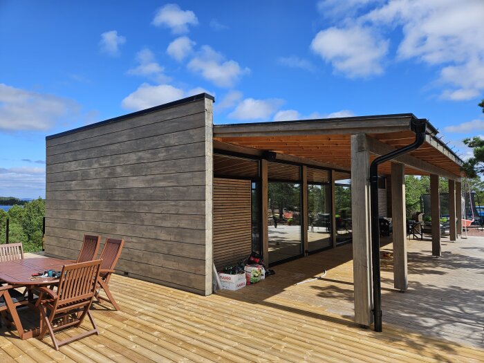 Uteplats med trädäck och veranda med glasväggar under klarblå himmel. Trädgårdsmöbler syns i förgrunden.