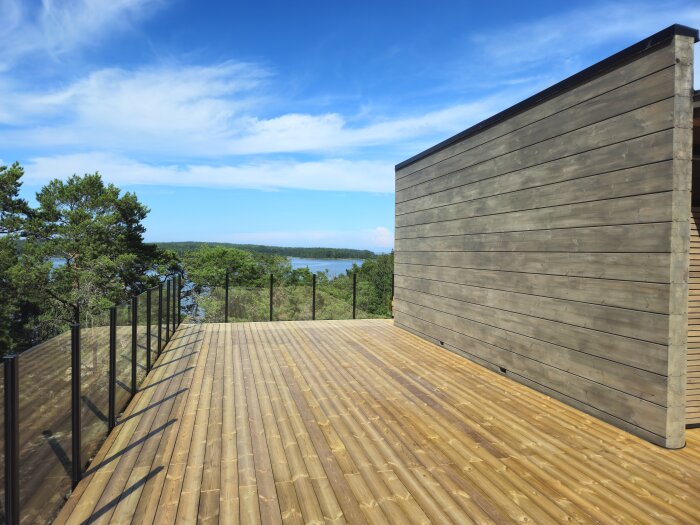 Trädäck med glasräcke och trävägg, utsikt över grönskande natur och sjö i bakgrunden under klarblå himmel.