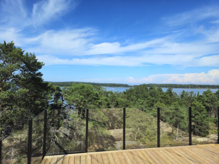 Trädäck med glasräcke och utsikt över skogslandskap och sjö under klarblå himmel, visar avslutat altanprojekt.