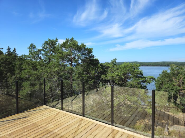 Träterrass med glasräcke och utsikt över skog och sjö under klarblå himmel.