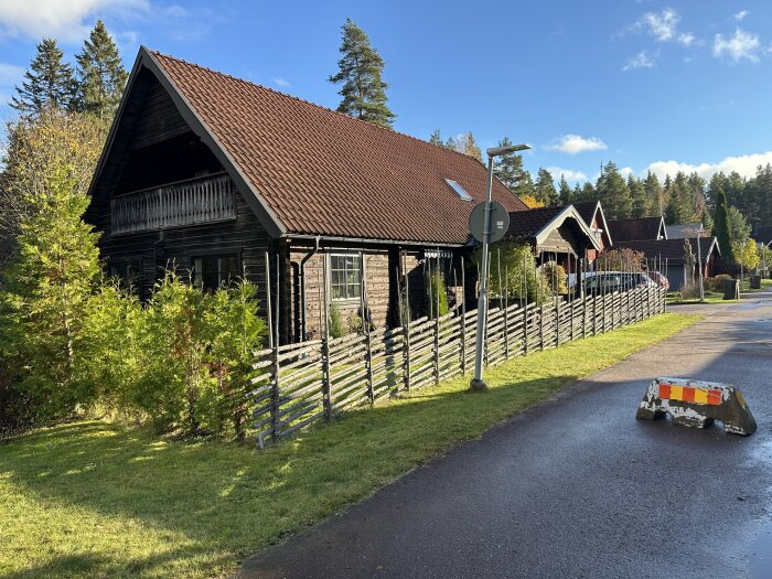 Timmerhus med brunt tak och järnvitriolbehandlad fasad, inhägnat med trästaket, omgivna av grönt landskap och fler byggnader i bakgrunden.