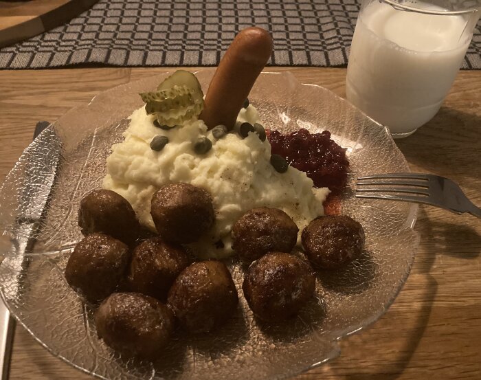 Köttbullar, potatismos med korv, lingon och kapris på en tallrik. Glas mjölk bredvid.