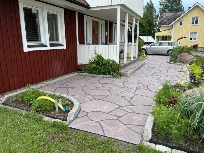 Gångväg av kalksten och kantsten vid rött hus med vit veranda, omgiven av grönska. Silverfärgad bil i bakgrunden.