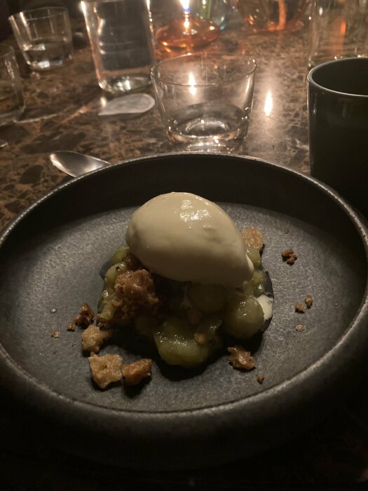 Efterrätt på tallrik med glasskula ovanpå karamelliserad frukt och smulor, placerad på ett marmorbord med vattenglas och ljus i bakgrunden.