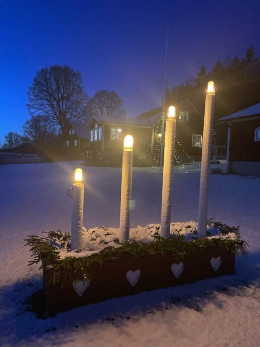 Dekoration med fyra flammande glödlampor som ser ut som levande ljus i en snötäckt trädgård, med bakgrund av belyst hus och träd.