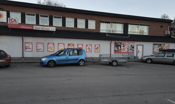 Blå bil med släpvagn parkerad utanför en ICA-butik i en tegelbyggnad med affischer på väggen.