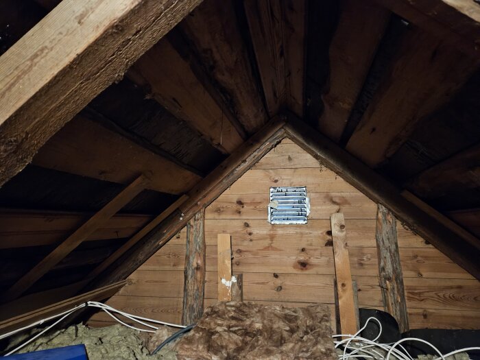 En vind i ett gammalt trähus med synliga träbjälkar och brädor. Det finns tecken på fukt och en ventilationsöppning i mitten av väggen.