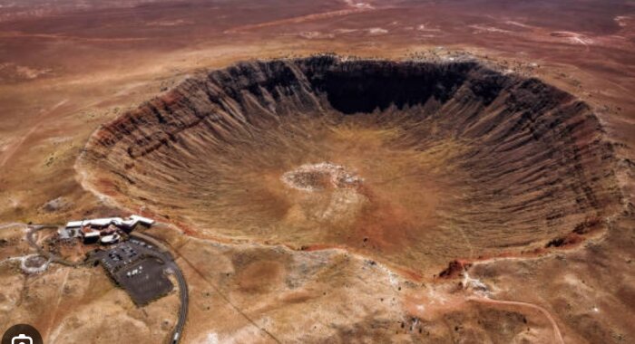 Flygfoto av en stor meteoritkrater i ett ökenlandskap med byggnader nära kraterkanten.