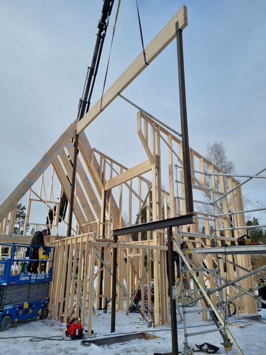 Byggarbetsplats med lyftkran monterar limträ och stålpelare på ett hus med synliga takstolar, snö på marken.