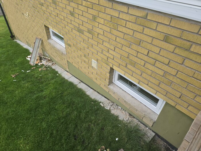 Nya fönster installerade i gul tegelvägg med rester av blåbetong och borttagna gamla fönster, liggande bredvid på gräsmattan.