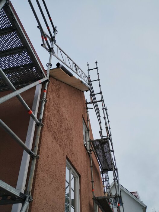 Byggnadsställning runt husets fasad med pågående arbete på takfoten; takstolar syns och är kapade vertikalt. Huset är i puts och ställningen i metall.