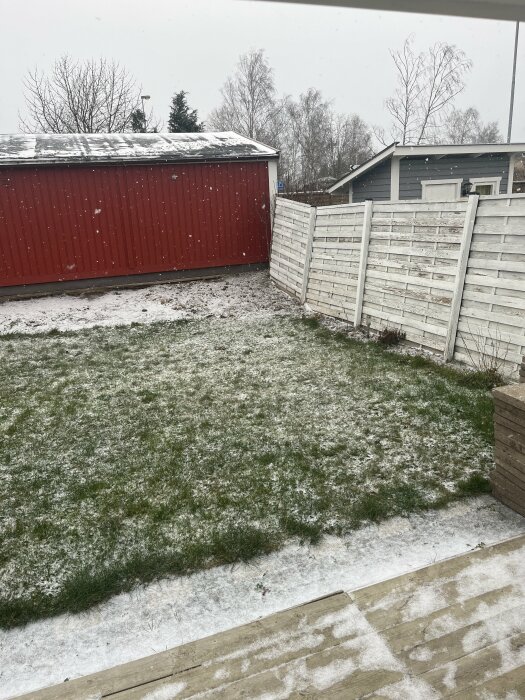 Snötäckt gräs och en vit, lutande träplanka bredvid en röd garagevägg med snö på taket.