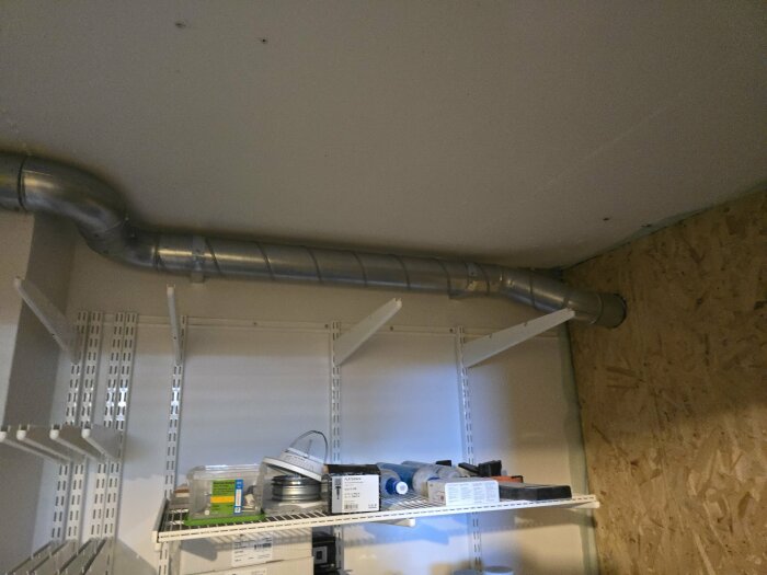 Ventilationsrör i maskinrum, rak installation i taket. Omgiven av hyllor med verktyg och material. Vägg med träskiva till höger.