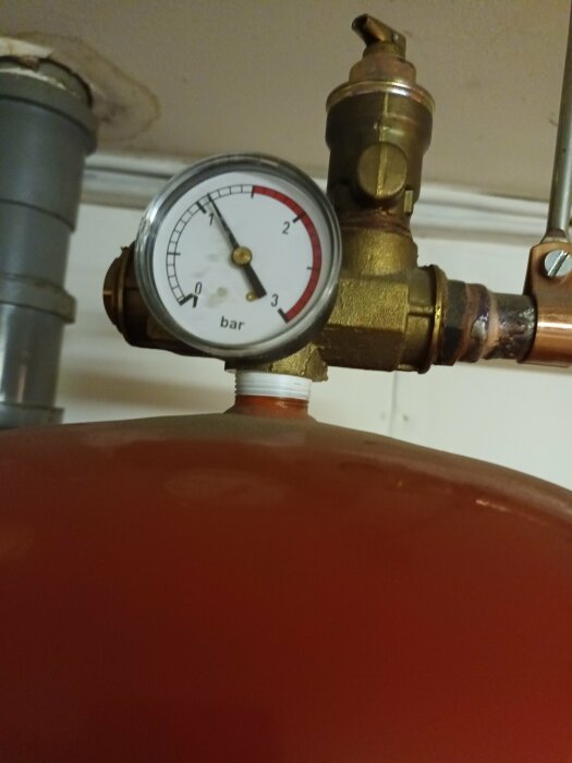 Manometer visar tryck på expansionstank i värmesystem, pekar på 1,2 bar. Kopparrör och del av röd tank synliga.