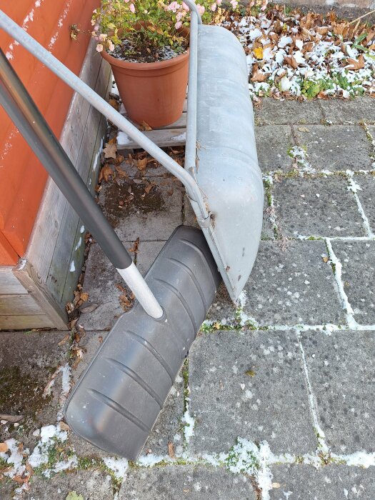 Snöskyffel och plogkäpp utanför ett hus, med höstlöv och lite snö på marken. En blomkruka syns bredvid.