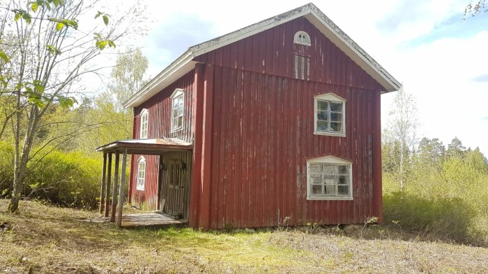 Röd gammal dalslandsstuga från 1800-talet i behov av renovering, omgiven av träd och buskage, med sprucken färg och gammeldags fönster.