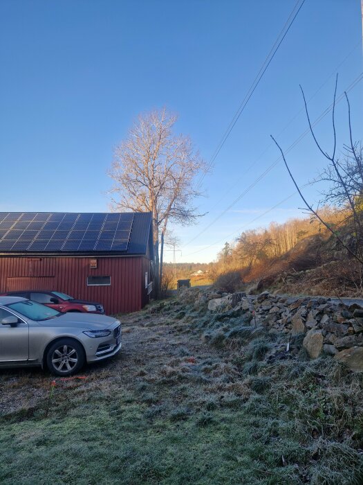 Hus med röda väggar och solpaneler på taket. Kraftledning i bakgrunden nära fastigheten. Bilparkeringsplats med två bilar framför huset.