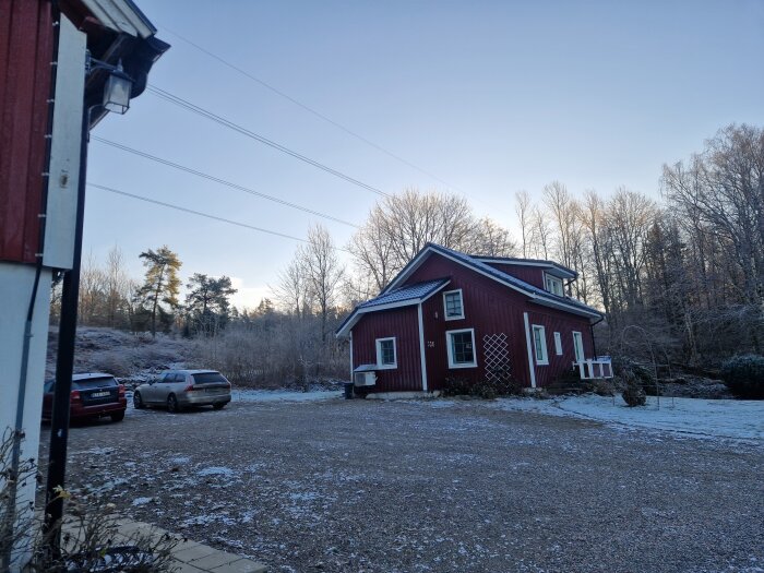 Röd byggnad med snötäckt mark, två bilar parkerade intill. Skogsbeklädd bakgrund under klar himmel.