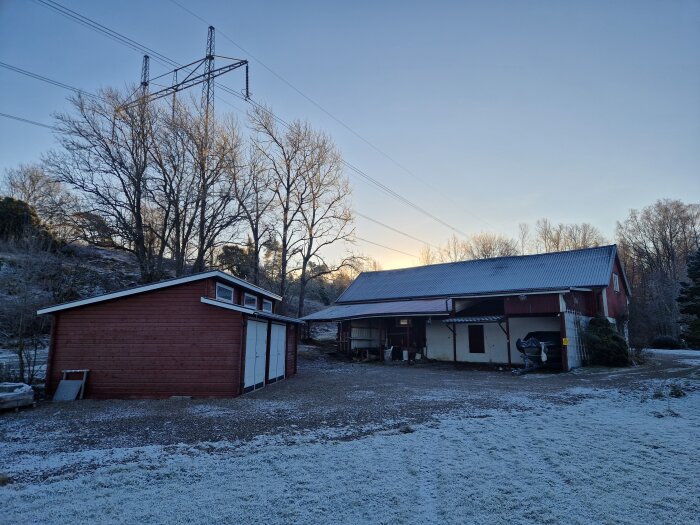 Röd ladugård och garage i vinterlandskap, träd och kraftledning i bakgrunden.