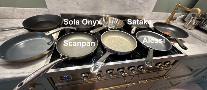 Fyra stekpannor på spisen: Sola Onyx, Satake, Scanpan och Alessi. Perspektivet gör de främre pannorna större.