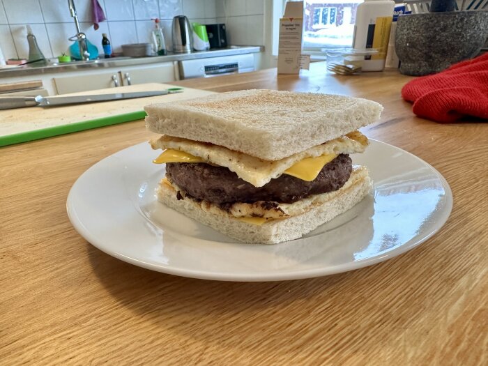 Improviserad hamburgare med stekt ägg och ost mellan två brödskivor, serverad på en vit tallrik i ett kök.