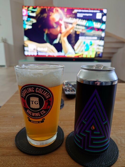 Ett glas öl från Toppling Goliath Brewing Co och en burk Omnipollo Maze med TV i bakgrunden.