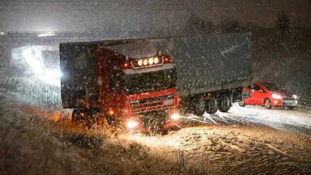 En långtradare fastnar i snöoväder på en hal väg, en röd bil syns i bakgrunden.