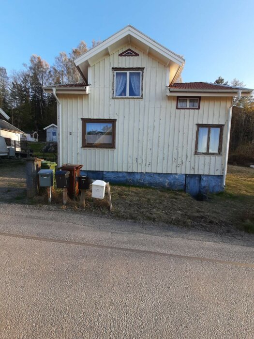 Gult trähus med blå sockel och röda takdetaljer, beläget vid en grusväg med postlådor framför. Skog i bakgrunden.