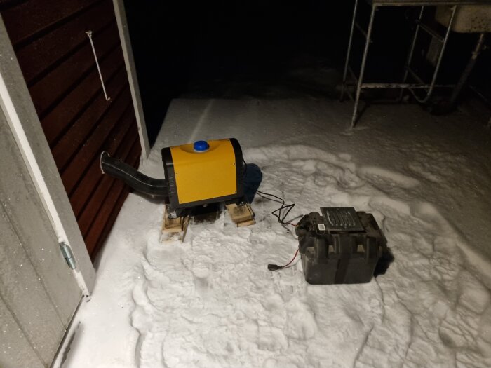 En värmeaggregat utomhus, placerat på snö vid en husvägg, med en kabel ansluten till ett batteri.