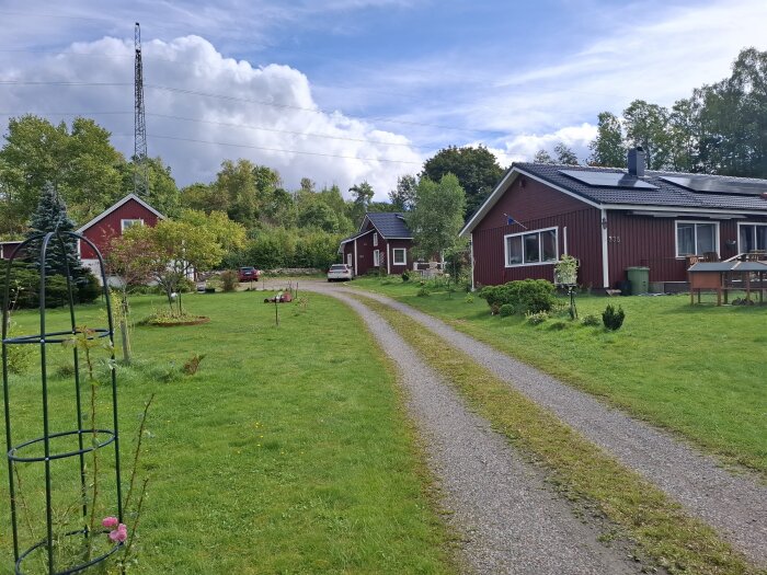 Vy av en grusväg med röda hus på sidorna, grönskande trädgårdar och en kraftledningsstolpe på en höjd i bakgrunden.