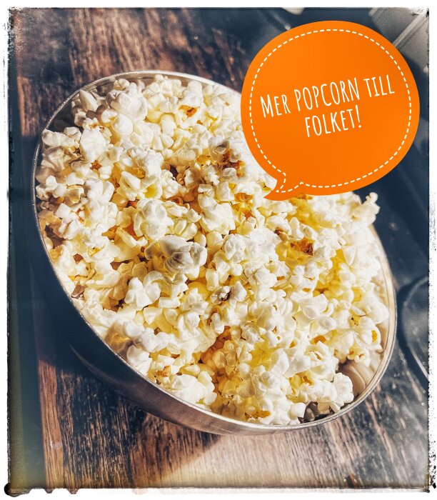 Skål med popcorn på ett träbord med pratbubbla: "Mer popcorn till folket!".