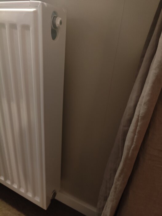 Vit radiator med synlig ventil upptill, mot en ljus vägg och med ett tygstycke bredvid. Frågeställning om ventilens placering för justering.