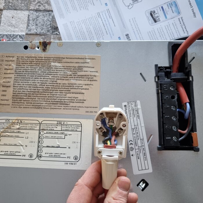 Närbild på baksidan av gammal spishäll med ansluten elkontakt som visar kabeldragning och anslutningsschema. Användarmanual synlig i bakgrunden.