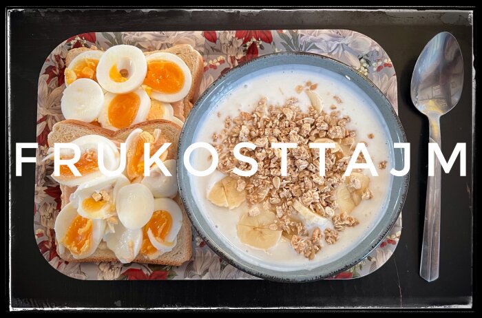 Äggmackor och skål med fil, banan och granola på bricka med texten "FRUKOSTTAJM", bredvid en sked på svart bord.