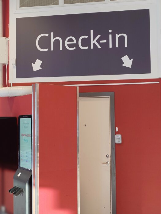 Självservice-incheckningsstation med skylt som visar "Check-in" och en dörr med kodlås i en byggnad med röda väggar.